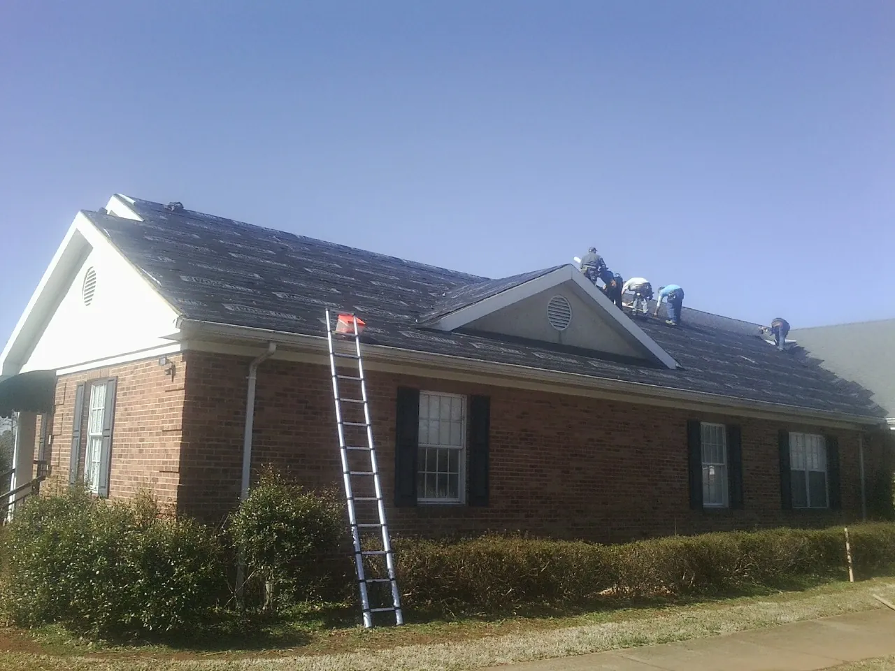 Crew tearing shingles off a brick ranch roof with a ladder set — Emergency Roof Repair work by our crew in Penfield