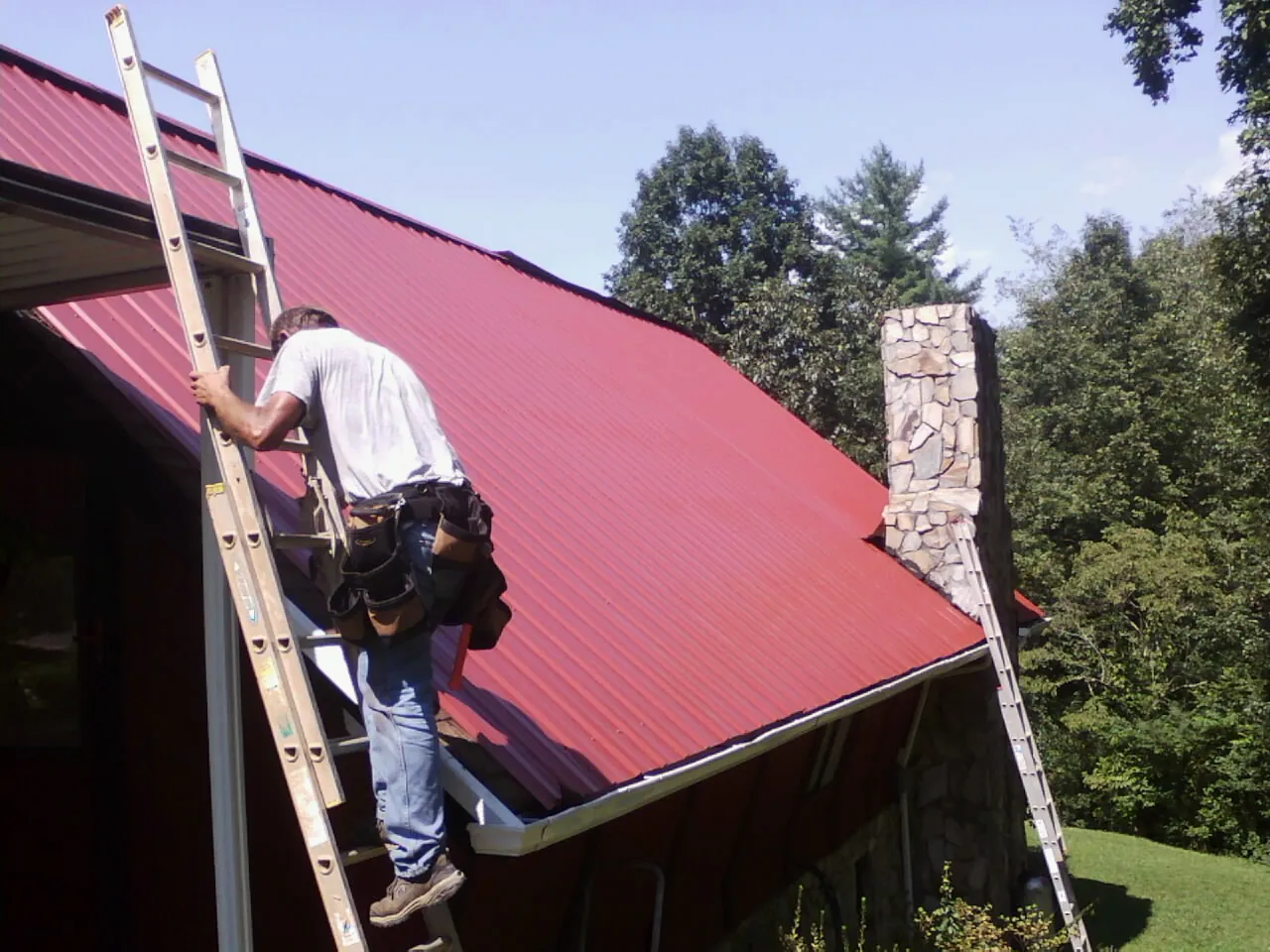 Roofer on a ladder beside a red metal roof and stone chimney — Soffit & Fascia Repair by Roofing Brothers in Penfield